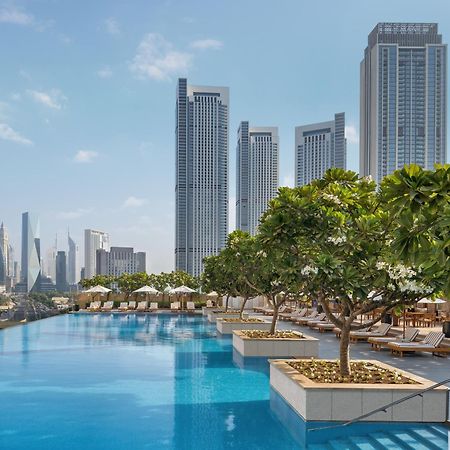 Kempinski Central Avenue Dubai Hotel Buitenkant foto The photo depicts a luxurious outdoor swimming pool area. The pool is surrounded by lush greenery and neatly arranged lounge chairs. In the background, there are tall skyscrapers, indicating an urban setting. The sky is clear with a few clouds, contr