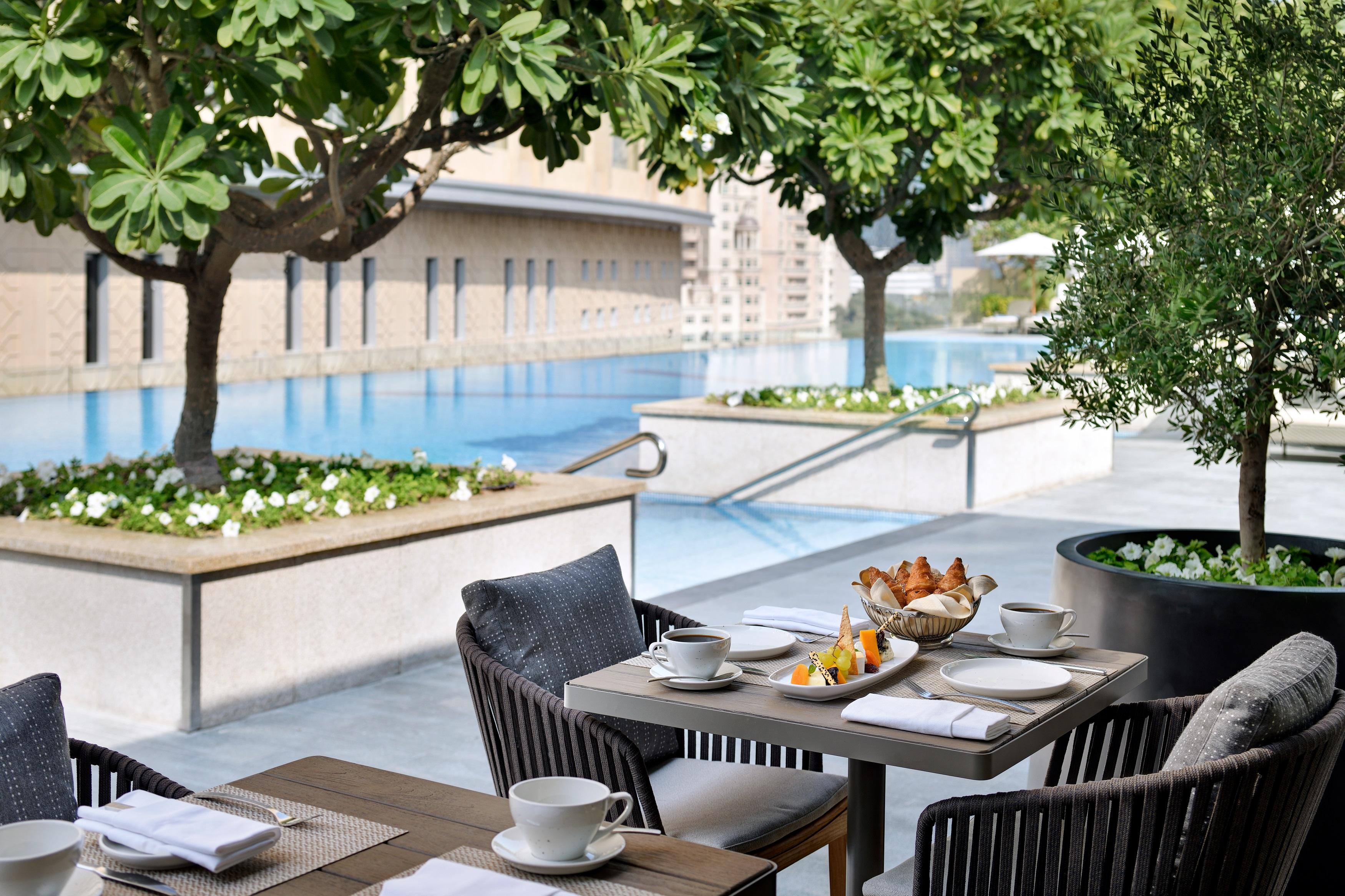 Kempinski Central Avenue Dubai Hotel Buitenkant foto The photo depicts a serene outdoor dining area beside a swimming pool. The table is elegantly set with cups, plates, and a variety of food, including pastries and fruit. Surrounding the area are lush green trees and decorative plants, enhancing the t