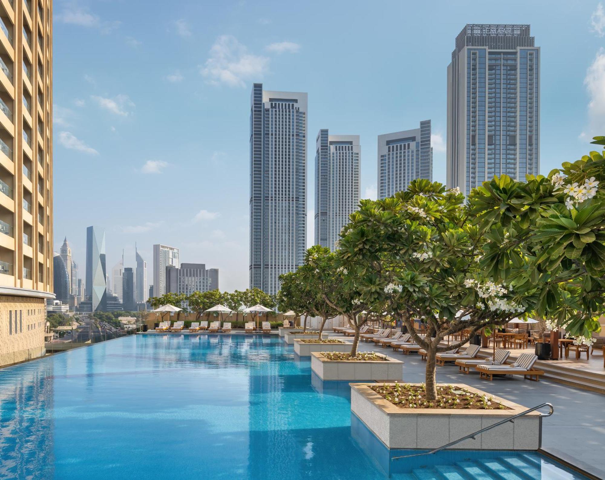 Kempinski Central Avenue Dubai Hotel Buitenkant foto The photo depicts a luxurious outdoor swimming pool area. The pool is surrounded by lush greenery and neatly arranged lounge chairs. In the background, there are tall skyscrapers, indicating an urban setting. The sky is clear with a few clouds, contr