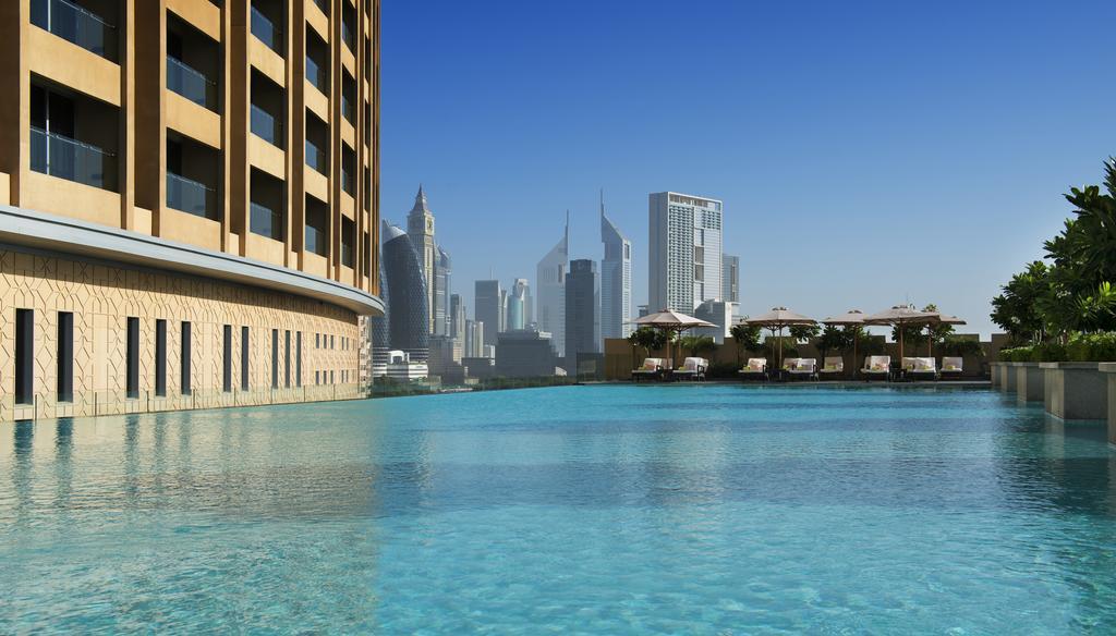 Kempinski Central Avenue Dubai Hotel Buitenkant foto The photo depicts a luxurious setting featuring a modern building with an elegant facade beside a large, clear blue swimming pool. In the background, a skyline of tall, contemporary skyscrapers is visible against a bright, sunny sky. The pool appears